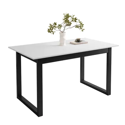 A white dining table with black U-shaped legs, decorated with a vase of green leaves and a book with a cup on top.
