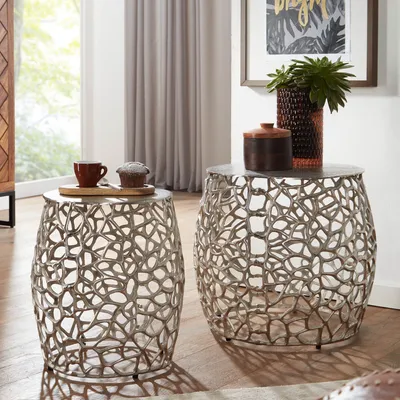 Two decorative side tables with intricate metal bases are shown in a living room setting. One table has a coffee cup and a muffin on it, while the other has decorative containers and a plant.