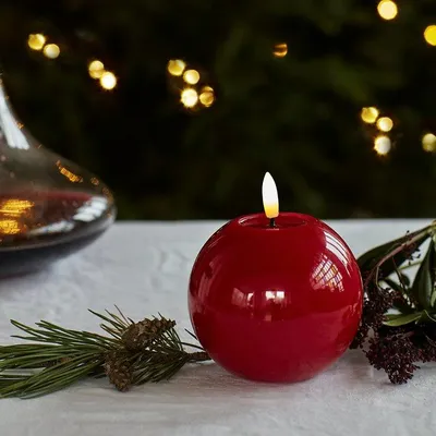 A red spherical LED candle with a flame is placed on a white tablecloth, with pine branches and pinecones in the foreground. In the background, out-of-focus Christmas lights are visible.