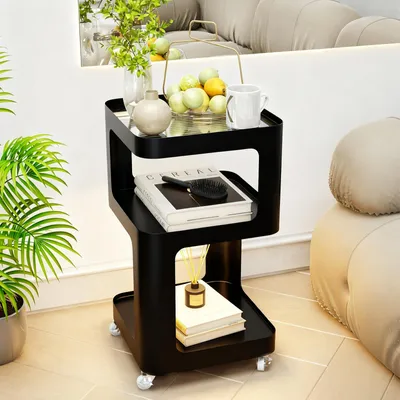 A black three-tiered side table with glass top, placed next to a sofa and a plant.