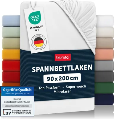 A white fitted sheet is displayed prominently, with a stack of other colored fitted sheets visible on the left and right sides. Several tags and certifications are attached to the sheet.
