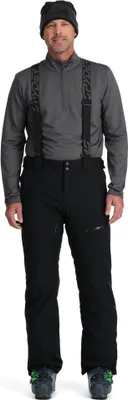 A man wearing ski gear, including a beanie, jacket, and pants with suspenders, stands facing the camera.