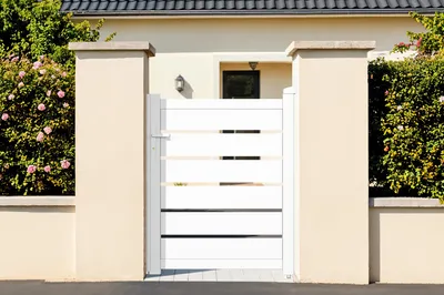 A white aluminum garden gate is shown in front of a house, flanked by beige pillars and lush green bushes.