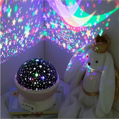 A star projector night light is casting colorful stars and moon shapes onto the walls and ceiling, illuminating a plush bunny toy wearing a crown.