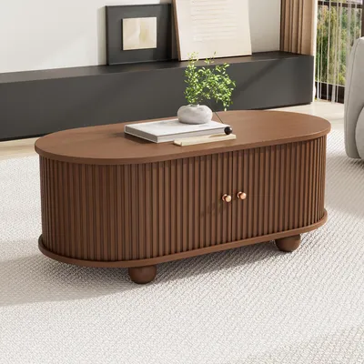 A wooden oval coffee table with ribbed doors and brass knobs sits on a textured rug in a living room setting.