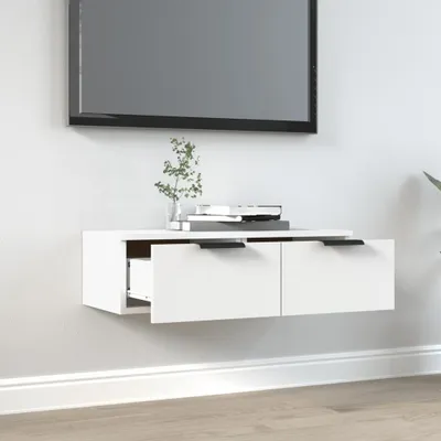 A white wall-mounted TV cabinet with two drawers, one of which is partially open, is shown mounted on a light gray wall above a white baseboard. A television is mounted on the wall above the cabinet, and a small plant in a vase and a stack of books are on 