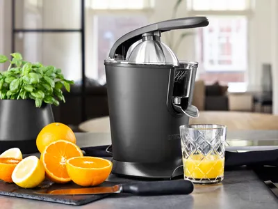 A black electric citrus juicer is shown on a kitchen counter next to cut oranges and lemons, and a glass of fresh orange juice.