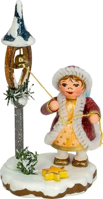 Ein kleines Kind in einem roten Mantel mit weißem Pelzbesatz steht auf einer schneebedeckten Basis und läutet eine goldene Glocke an einem dekorativen Ständer während es zwei gelbe Sterne neben sich hat und von Tannenzweigen umgeben ist