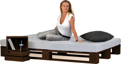 A woman sitting on a bed frame made of wooden pallets, with a nightstand next to it.