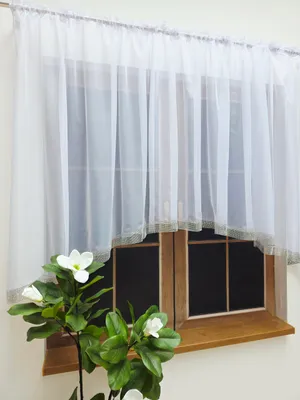 A white sheer curtain with a decorative border is hung over a wooden window. A plant with white flowers is in the foreground.