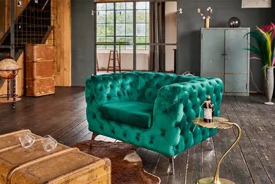 A green velvet armchair in a loft-style living room with wooden floors and industrial-style furniture.