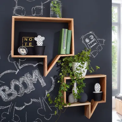 Two wooden shelves shaped like speech bubbles are mounted on a dark wall with chalk drawings. One shelf holds books and decorative items, while the other has plants and vases.