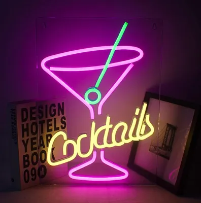 A neon sign shaped like a cocktail glass with the word 'Cocktails' below it, displayed in a room with books and a framed picture.
