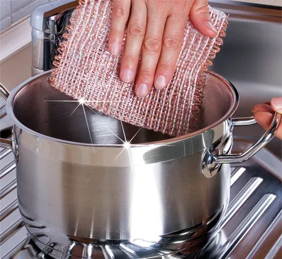 A hand is shown scrubbing the inside of a stainless steel pot with a copper-colored scrubber.