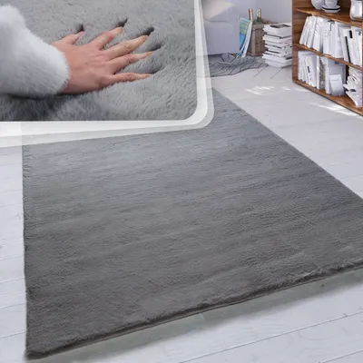 A hand pressing into a soft, gray, high-pile rug, showcasing its plushness and texture. The rug is laid out on a light-colored wooden floor, with a bookshelf and part of a living room visible in the background.