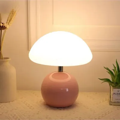 A pink mushroom-shaped table lamp with a white shade sits on a textured surface next to a vase of dried flowers and a small potted plant.