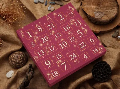 A close-up of a red advent calendar with gold numbers and festive designs, set against a textured brown burlap background with natural elements like stones and wood.