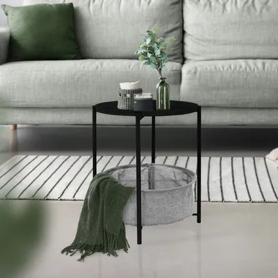 A round black metal side table with a gray fabric basket and a green blanket draped over it, placed on a striped rug next to a light gray sofa.
