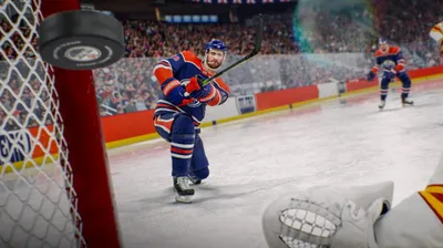 A hockey player in a blue and orange uniform is skating on an ice rink, about to hit a puck that is flying towards the camera. The background shows a stadium filled with spectators.