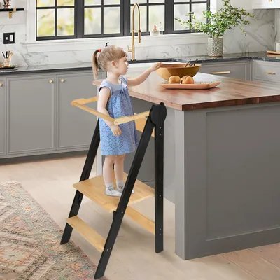 A young girl stands on a wooden step stool in a modern kitchen, reaching for a bowl on the counter.