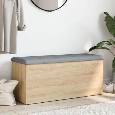 A storage bench with a gray upholstered top and a wood-effect body, placed in a hallway setting with a coat hanging on the wall and a plant in the background.