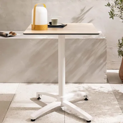 A white table with a wooden top and a white base, with a lamp and a cup on it, set on a tiled floor with a concrete wall in the background.