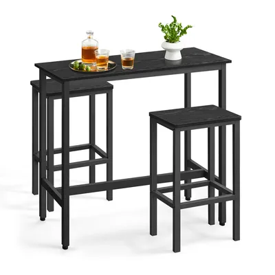 A black bar table with two black bar stools, set against a white background. There are two glasses of amber liquid and a bottle of amber liquid on the table, along with a small green plant in a white pot.