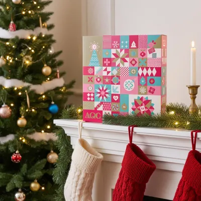 A Christmas-themed advent calendar box sits on a mantelpiece next to a decorated Christmas tree and stockings.