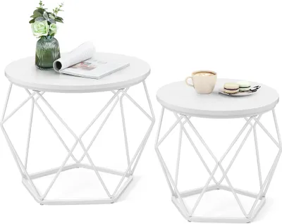 Two white geometric side tables are displayed against a white background. The larger table on the left has a vase with green flowers and a magazine on it. The smaller table on the right has a cup of coffee and macarons on it.