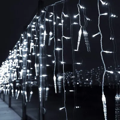 A string of icicle-shaped lights with white LEDs, hanging in front of a blurred cityscape at night.
