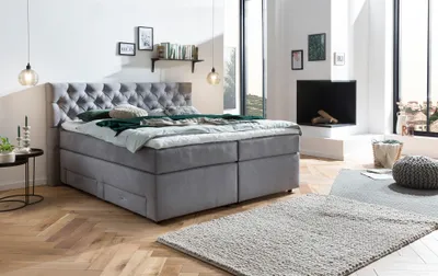 A bedroom scene with a large grey boxspring bed as the focal point, featuring a Chesterfield headboard and drawers. The room is decorated with a fireplace, rugs, and hanging lamps, suggesting a modern and cozy living space.