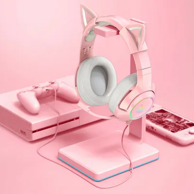 A pink gaming headset with cat ears is displayed on a pink stand, next to a pink gaming console and controller, and a pink smartphone.