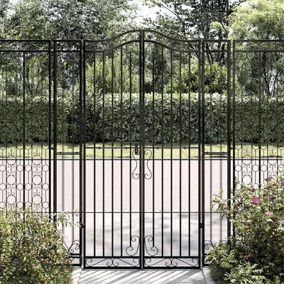 A black wrought iron garden gate with decorative scrollwork and a matching fence section, set against a backdrop of lush green bushes and trees.