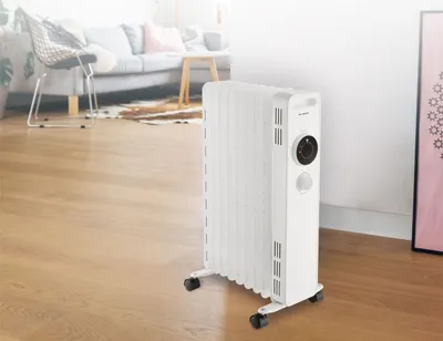 A white oil radiator is placed on a wooden floor in a living room setting, with a sofa and chair in the background.