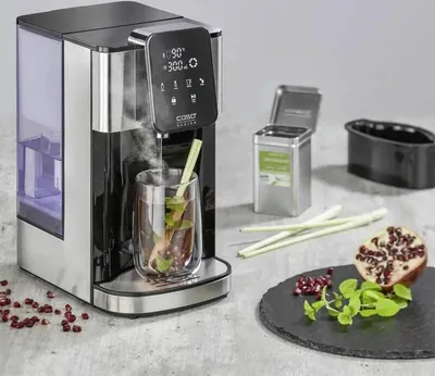 A CASO HW 1660 Turbo hot water dispenser is shown in use, dispensing hot water into a glass with tea leaves and herbs. Other items like a tea tin, pomegranate, and mint leaves are also present.