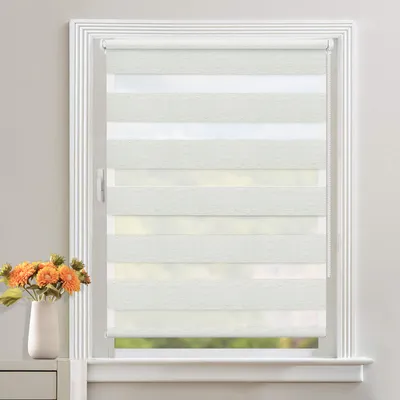 A white roller blind with horizontal stripes of opaque and sheer fabric is installed in a white window frame. A vase with orange flowers is placed on a shelf to the left of the window.