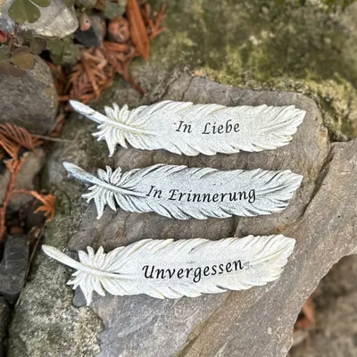 Three decorative feather-shaped grave decorations with German inscriptions are placed on rocks and foliage.