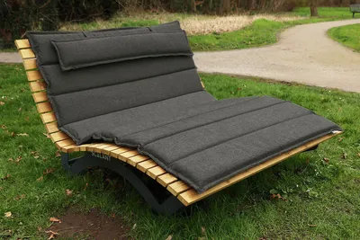 A wooden chaise lounge with a dark gray cushion sits on a grassy area next to a paved path in a park.