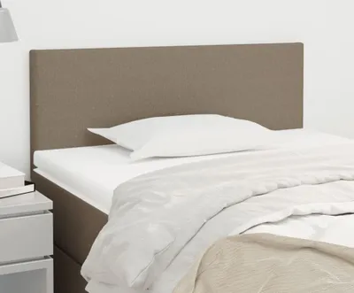 A close-up view of a taupe fabric headboard attached to a bed frame, with a white pillow and duvet.