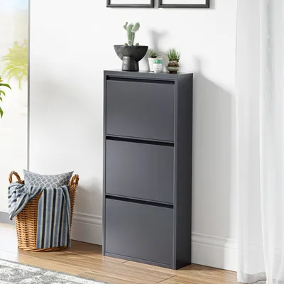 A dark gray shoe cabinet with three flip-down doors, decorated with small potted plants on top, stands against a white wall next to a window and a wicker basket filled with blankets.
