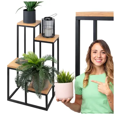 A woman is pointing to a multi-tiered plant stand with plants and a lantern on it. The woman is holding a pink pot with a plant.