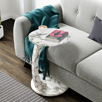 A marble-look C-shaped side table with a book and coffee cup on it, placed next to a gray sofa with a teal blanket draped over it.