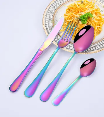 A set of rainbow-colored cutlery is arranged on a white surface, with a plate of spaghetti in the background.