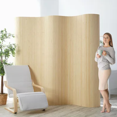 A woman stands holding a mug next to a room divider, with a chair and plant in the foreground.