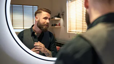 A man with a beard is looking in a mirror and adjusting his hair and beard.