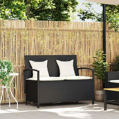 A black poly rattan garden bench with white cushions is placed on a patio in front of a bamboo fence. There is a small white table with a plant to the left of the bench, and a potted plant to the right.