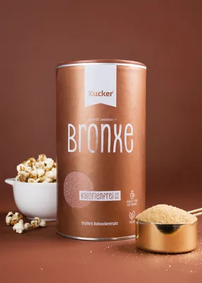 A bronze colored canister of Xucker Bronxe Erythrit sweetener is shown in the center, with a bowl of popcorn to the left and a measuring cup filled with the sweetener to the right.