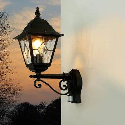An outdoor wall lamp with a motion sensor is mounted on a white wall, with a sunset sky and trees in the background.