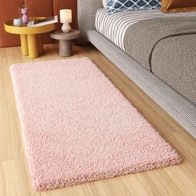 A pink rug is placed on a wooden floor next to a bed and a small side table with decorative items.