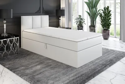 A white boxspring bed with a headboard is shown in a room with a large rug and plants.